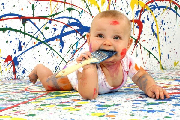 Piccolo bambino coperto di vernice che lecca con un pennello sorridente