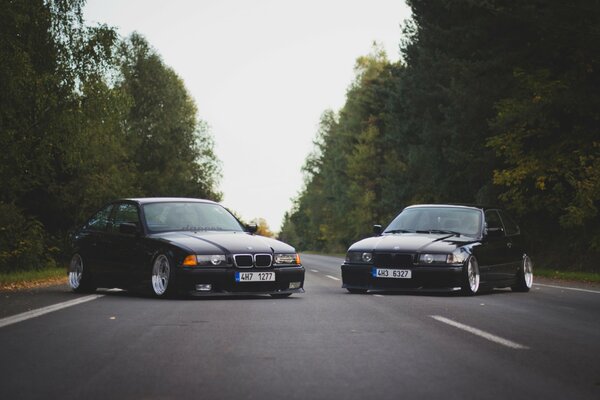 BMW M3 e36 3 series oldschool tuned cars stand across an asphalt road against a forest background front view