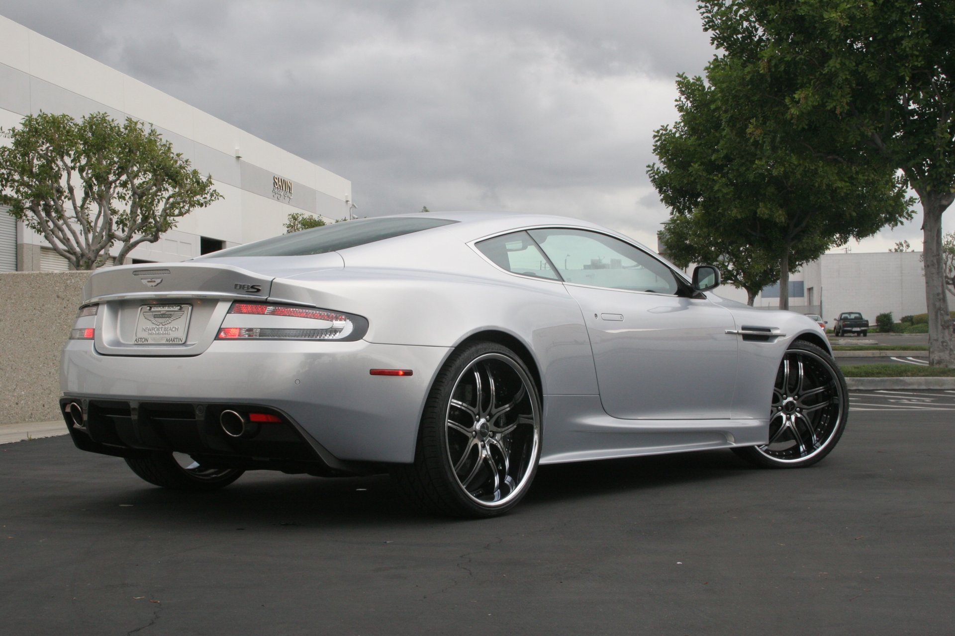 aston martin dbs plateado ruedas aston martin dbs plata vista trasera luz de faro cielo nubes árboles edificio