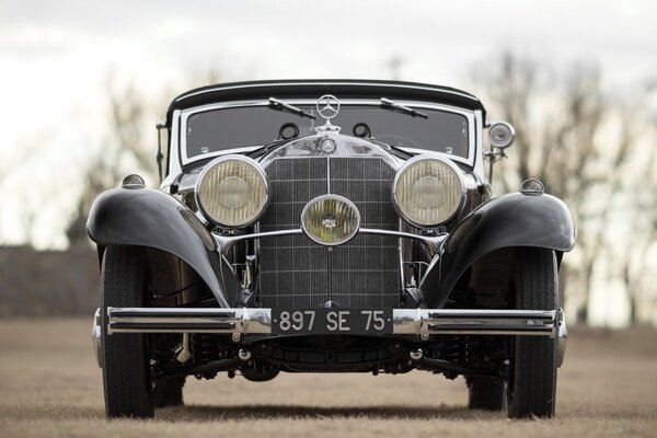 A black Mercedes-Benz 540k stands on withered grass in an open space