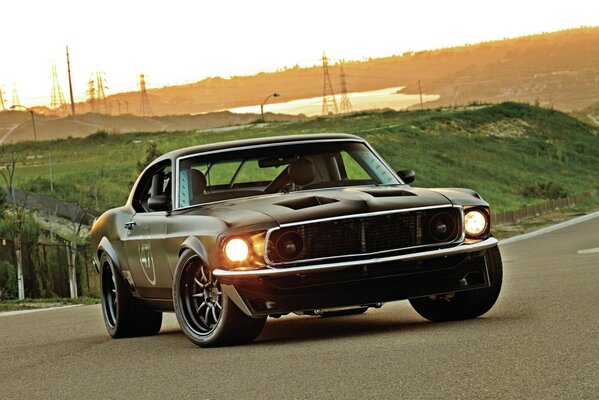 Black Ford Mustang with burning headlights on sunset background