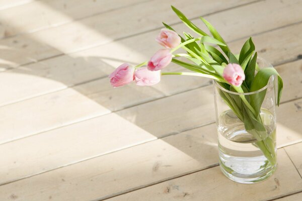 Tulipanes en un vaso de agua en un piso de madera bajo el sol de la mañana de primavera