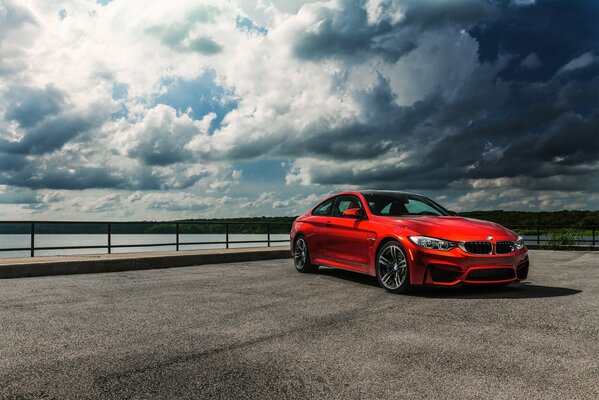 A bright scarlet stylish car stands on a bridge over a river under beautiful clouds