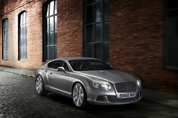 A grey Bentley Continental car stands next to a brick factory