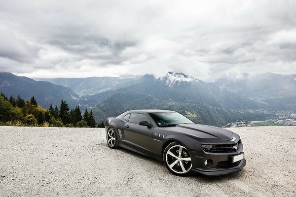 Voiture noire Camaro Chevrolet sur la route sur fond de forêt de montagnes et ciel nuageux