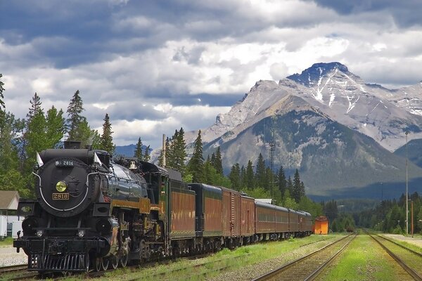 A steam locomotive at a station in the mountains under a cloudy sky