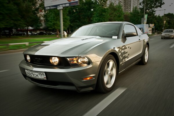 On the highway, a silver Mustang car with its headlights on