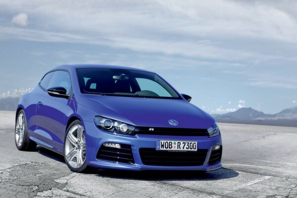 Ein VW Scirocco blau auf dem Hintergrund der blauen Berge unter einem bewölkten Himmel