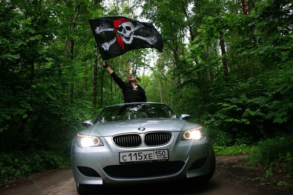 Mężczyzna z piracką flagą na szarym BMW na leśnej drodze