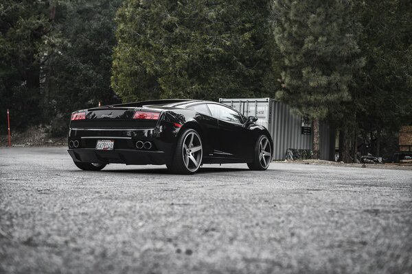 Coche negro marca Lamborghini vista trasera lateral sobre el fondo de asfalto y árboles