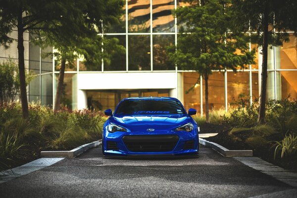 A blue Subaru stands in the shade of bushes, next to a beautiful building