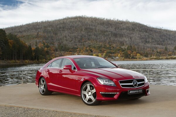 Cielo, montaña, bosque y sedán rojo de Mercedes CLS63 en la orilla del lago