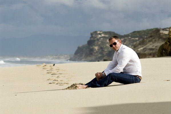 Daniel Craig w ciemnych okularach siedzi na plaży boso