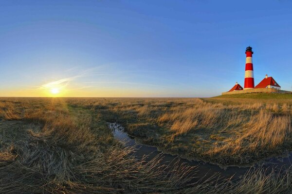 Ein Panorama-Morgen öffnet den Blick auf das Grasfeld, ein fließender Bach und ein Leuchtturm steht in der Ferne