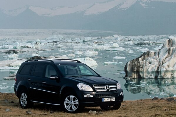 Black Mercedes SUV on the shore of a pond with melting ice and mountains in the distance