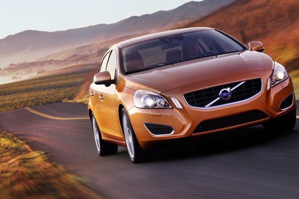 An orange Volvo car on the road in a field against the background of mountains and clear blue sky