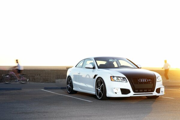 There is a white Audi in the parking lot, and a cyclist in the background