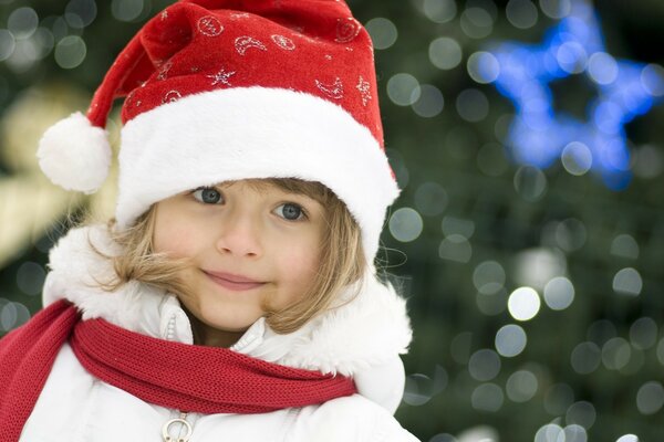 Ein Mädchen mit einem Quadrat in einer Neujahrsmütze. Ein blauäugiges Mädchen in einem Weihnachtsmann-Hut. Neujahrsfoto mit Kindern. Mädchen auf einem Weihnachtsbaum Hintergrund