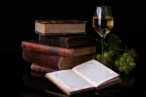 Pila de libros, libro abierto, vino y uvas verdes en la mesa