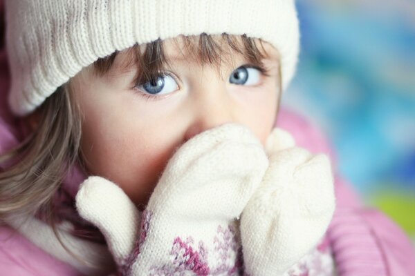 A blue-eyed girl covers her face with her hands in warm white mittens