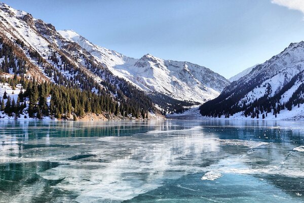 Frozen lake, mountains covered with snow, forest on the slopes of the mountains. Sunny day