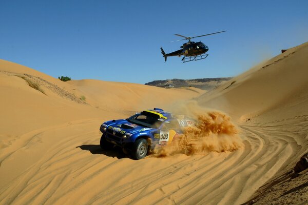 A blue SUV is driving on the sand and a helicopter is flying behind it