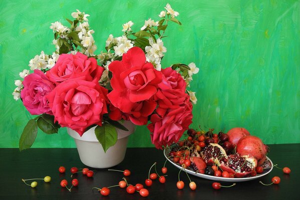 On the table there is a pot of roses and a plate of pomegranates and cherries