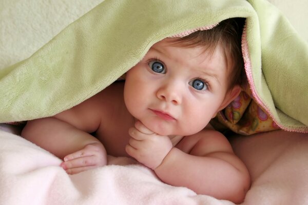 The baby looks out from under the green blanket