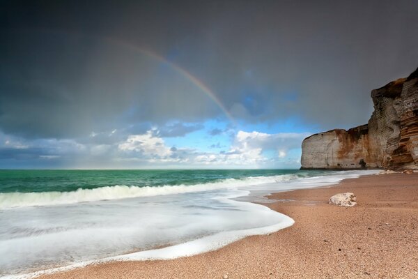 Mare verde, surf schiumoso e spiaggia sabbiosa con roccia sullo sfondo di un arcobaleno nebbioso