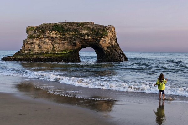 The girl is standing on the sand next to a rock