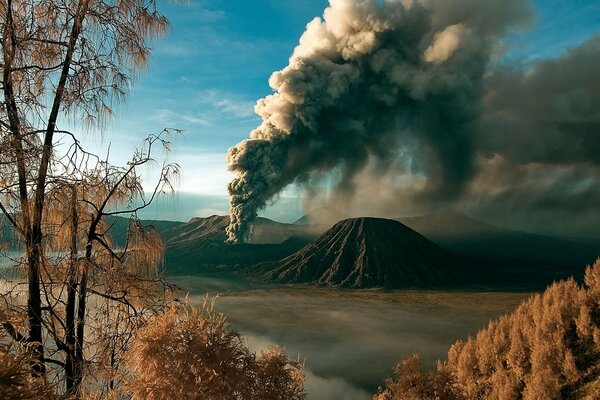 Paisaje con un volcán y nubes de humo en el fondo de un bosque otoñal y un cielo azul brillante