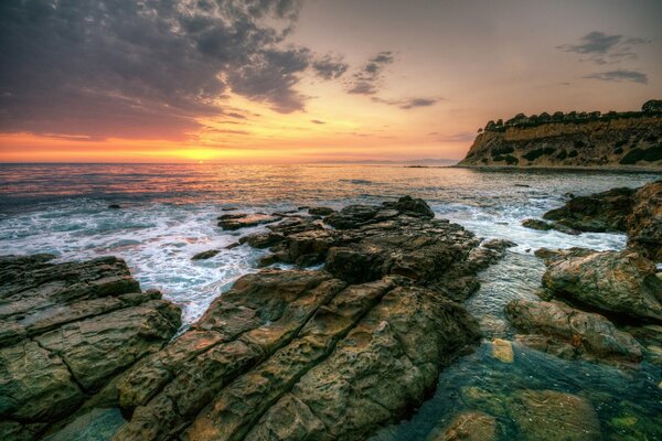 Costa rocosa junto a las olas de espuma teñidas con una franja amarilla-naranja de puesta de sol cubierta de nubes marrones