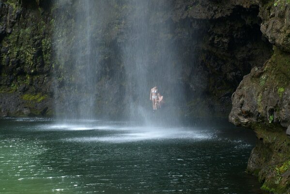 A couple is sitting under a rock and a waterfall flows nearby