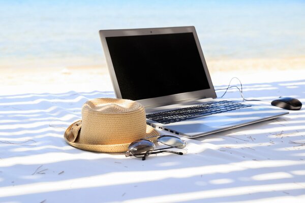 Ordinateur portable et chapeau se trouvent sur le sable blanc