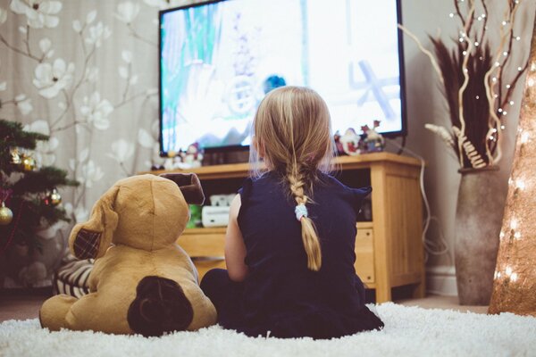 Kleines Mädchen mit einem Spielzeug auf dem Bett, das Silvester fernsieht