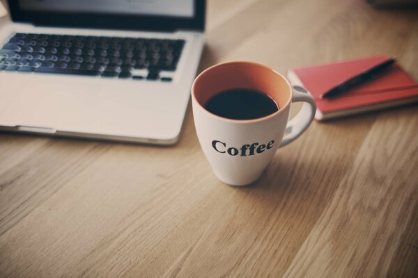 A cup of coffee, a white laptop and a red notebook on a wooden table