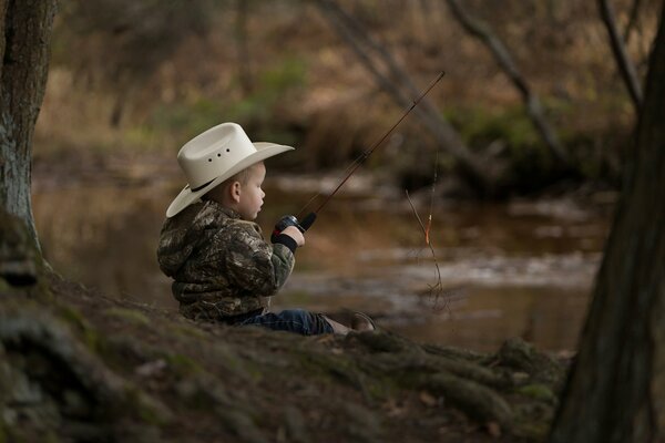 A kid in a hat with a fishing rod is sitting on the shore