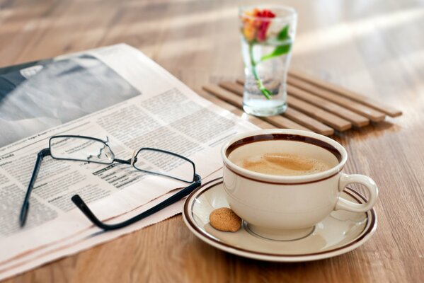 Eine Tasse Kaffee mit einem kleinen Keks steht auf dem Tisch, daneben befinden sich eine Zeitung, eine Brille und ein Glas mit einer Blume