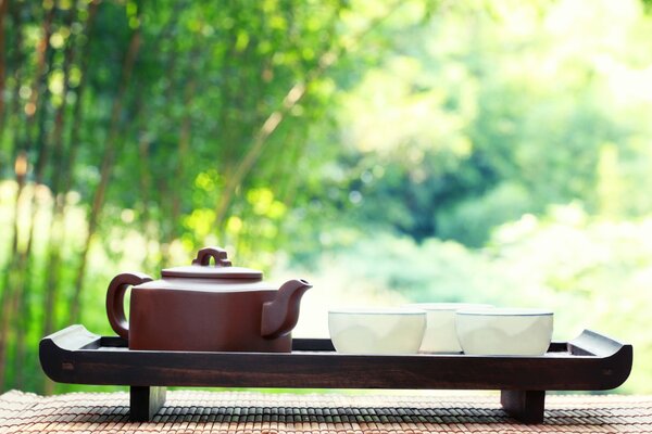 A brown teapot and three white cups stand on a tray against the background of nature