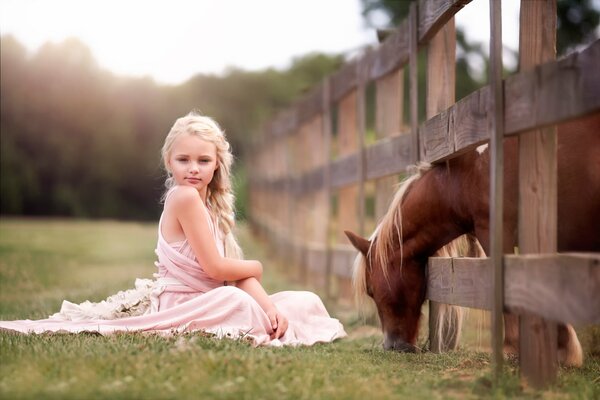 A funny girl is sitting on the grass, and a horse is grazing behind the fence