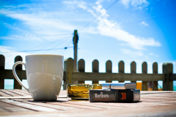 Taza, cenicero, encendedor y cigarrillos contra el cielo azul