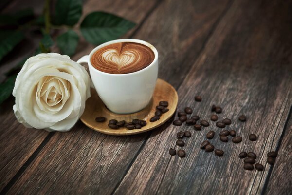 Rose blanche sur la table avec une tasse blanche remplie de café en mousse avec des grains de café parfumés à côté