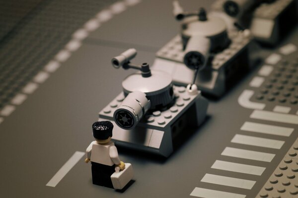 A man in a white shirt, black trousers and with a white briefcase stands face to face with gray tanks, all made of Lego