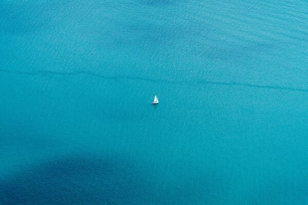 Lontano dalla riva sul mare blu galleggia una barca e solo una vela bianca è visibile