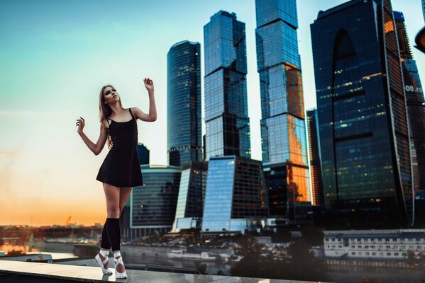 Ein Mädchen in einem schwarzen Kleid und weißen Ballettschuhen steht vor dem Hintergrund von Moskau City