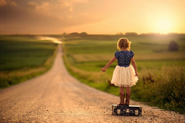 The girl is standing on a suitcase on the road