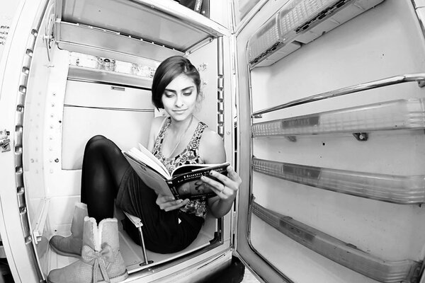 A fragile girl is sitting in the refrigerator and reading a book