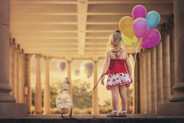 A little girl in a dress with berries holds a goose and colorful balloons on a red leash