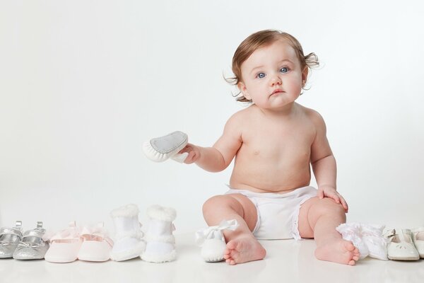 Kleines Baby sitzt auf dem Boden in einer Windel neben kleinen Schuhpaaren von Schuhen