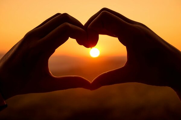 A girl s hands in the shape of a heart on the background of a sunset